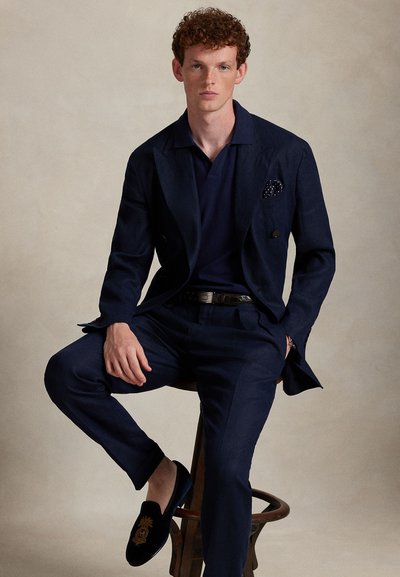 Navy suit with double-breasted jacket and matching trousers, textured fabric, paired with a navy polo shirt and black loafer-style shoes.