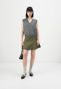 Woman wearing a gray sleeveless V-neck sweater, olive green skirt, beige socks, black pointed shoes, and holding a small black handbag.