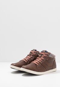 Brązowe skórzane sneakersy w stylu high-top z różowymi sznurowadłami, teksturowanym kołnierzem, białą gumową podeszwą oraz subtelnym logotypem z boku.