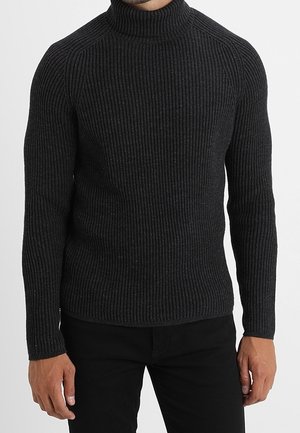 Homme portant un pull à col roulé côtelé noir ajusté et un pantalon noir, debout les bras détendus le long du corps.