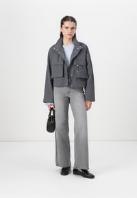 Kort grå jakke med to frontlommer, sølvfarget detaljer og høy krage, kombinert med vide grå jeans og en svart håndveske.