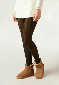 Leggings lavorati a maglia marroni abbinati a stivali alla caviglia color cammello con una soffice fodera e una punta rotonda. L'outfit è completato da una tunica bianca.