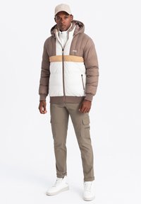 Chaqueta acolchada con un diseño de bloques de color marrón, blanco y crema; cuenta con capucha, bolsillos con cremallera y puños de canalé. Combinada con pantalones cargo.