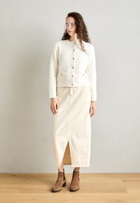 Cárdigan blanco de punto con botones dorados, combinado con una falda midi beige con una abertura frontal y botas de tobillo marrones.