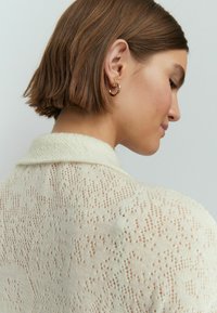 Cardigan a maglia in morbido crema con un motivo in pizzo testurizzato, colletto a punta e dettagli in tessitura aperta. Orecchino a cerchio dorato visibile.