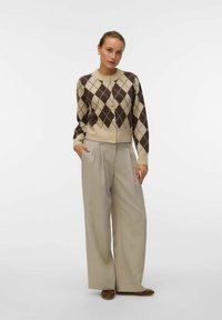 Cardigan beige con motivo argyle e diamanti marroni, abbottonatura frontale e polsini a coste. Abbinato a pantaloni larghi grigi chiari e scarpe marroni.