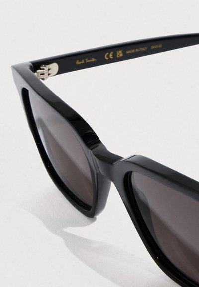 Μαύρα γυαλιά ηλίου με γεωμετρικό σχήμα cat-eye, με σκουρόχρωμους φακούς και γυαλιστερά πλαστικά πλαίσια. Μεταλλικά στοιχεία κοντά στις μεντεσέδες.
