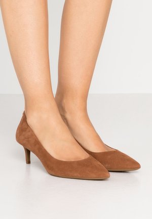 Pieds chaussés de chaussures en daim marron à bouts pointus et talons kitten, debout sur un sol de couleur claire.