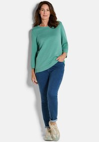 Hellblauer Pullover mit strukturiertem Muster, runder Halslinie und drei Viertelärmeln. Kombiniert mit blauen Jeans und beigen Sneakers.