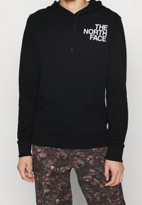 Svart hoodie med fram-känguruficka, justerbar dragsko och vit "THE NORTH FACE"-logga på vänster sida. Kombinerad med mönstrade byxor.