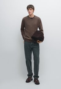 Brown knit sweater, dark gray jeans, brown leather shoes. Model holding a dark brown hat. Simple background, neutral colors.