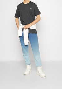 Person wearing dark grey Nike t-shirt, ombre blue joggers, white sneakers, and white sweatshirt tied around waist on plain background.