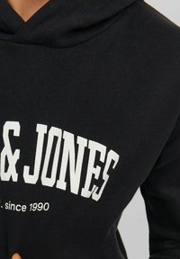 Sudadera negra hecha de una mezcla de algodón, con un gran logo impreso en blanco "JONES" y el texto "desde 1990" en una fuente blanca más pequeña.