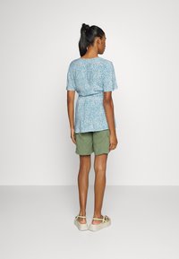 Top azul de manga curta com padrões brancos, cintura ajustada, combinado com calções verdes e sapatos claros com tiras verdes.