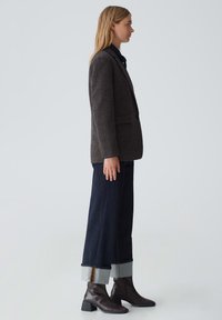 Blazer marrone texturizzato, jeans scuri a gamba larga con risvolti grigi, e stivali con tacco marroni. Linee pulite, vestibilità su misura, design minimalista.