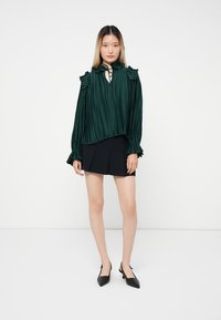 Blusa verde a pieghe con spalle arricciate e dettagli al collo con fiocco, abbinata a una gonna nera e scarpe nere a punta.