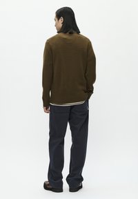 Man with shoulder-length black hair wears brown sweater, dark loose pants, and brown shoes, standing with back facing camera.