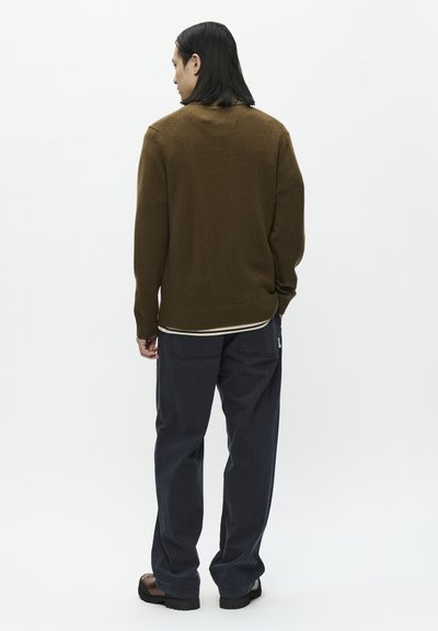 Homme aux cheveux noirs mi-longs portant un pull marron, un pantalon large sombre et des chaussures marron, se tenant de dos par rapport à l'appareil photo.