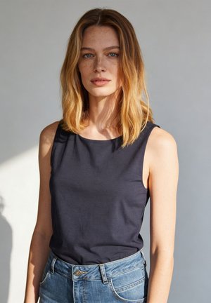 Young woman with shoulder-length blonde hair, freckles, wearing a sleeveless black top and blue jeans, standing against a plain light background.
