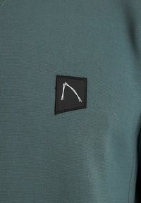 Sudadera verde con una tela texturizada y un parche rectangular negro que muestra líneas abstractas blancas. Diseño simple y moderno.