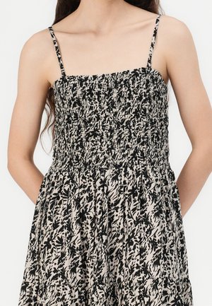 Femme portant une robe sans manches avec de fines bretelles et un tissu à motif floral noir et beige. Mains reposant derrière son dos.