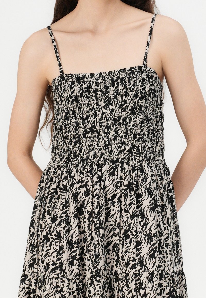 Femme portant une robe sans manches avec de fines bretelles et un tissu à motif floral noir et beige. Mains reposant derrière son dos.