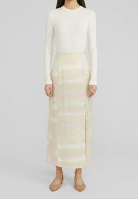 Kvinna iförd en vit långärmad ribbad topp, en pastellfärgad tie-dye midi kjol med slits på sidan och vävda spetsiga ballerino skor, ståendes mot en neutral bakgrund.