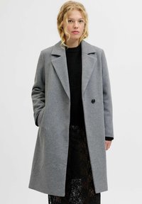 Manteau en laine gris avec un col cranté, fermeture à un bouton, et poches latérales, porté sur un pull en maille noir et une jupe en dentelle.