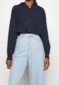 Námořnická modrá košile s dlouhým rukávem a špičatým límcem, zapínání na knoflíky a volný střih, spárovaná se světle modrými džíny s vysokým pasem.