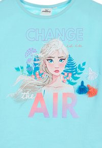 Camiseta de color azul claro con un gráfico de un personaje de cabello largo, el texto "EL CAMBIO está en el AIRE" y borlas decorativas coloridas.