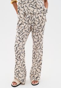 Patroon, lichte beige broek met zwarte draaiende ontwerpen. Heeft een losse pasvorm en flared pijpen, gecombineerd met beige slippers.