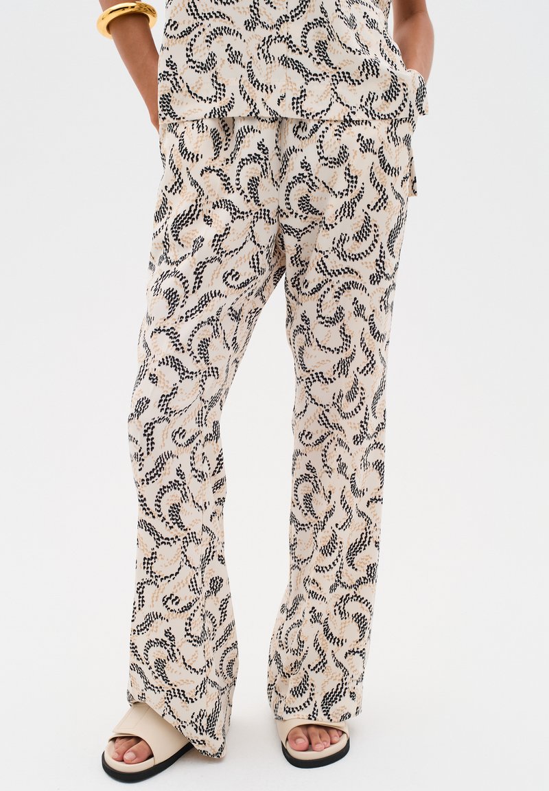 Pantalons beiges légers à motifs avec des designs noirs en spirale. Présentent une coupe ample et des jambes évasées, associés à des sandales beiges.