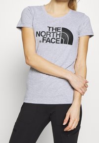 Osoba v krátkém šedém tričku s krátkým rukávem a černým logem "THE NORTH FACE" a černých kalhotách, která stojí s překříženýma rukama vpředu.