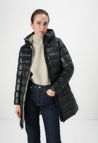 Čierny puffa kabát s kapucňou, lesklý povrch, zips vpredu a prešívaný dizajn. Nosený cez krémový sveter a tmavé džínsy.