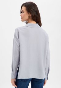 Light gray long-sleeve shirt with a collar, featuring a relaxed fit, cuffed sleeves, and a slightly curved hem. Smooth, textured fabric.