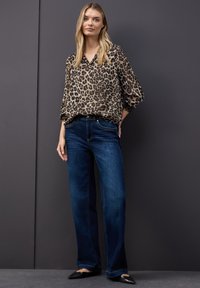 Blusa stampa leopardata con scollo a V e maniche a sbuffo abbinata a jeans a gamba larga blu scuro e ballerine nere con punta appuntita su sfondo grigio.