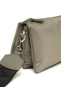 Wypchana oliwkowa torba z teksturowanym paskiem, zamknięciem na zamek błyskawiczny i srebrnymi akcesoriami. Ma szczegółowy akcent z materiału w dopasowanym kolorze.