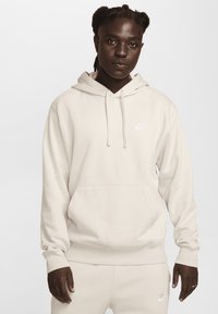 Bežs kapučjaka krekls ar auklēm, priekšējo kabatu un mazu baltu Nike logo uz krūts. Audums izskatās mīksts un gluds.