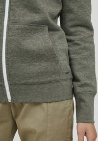 Haut à capuche zippé gris avec poches avant, fabriqué à partir d'un tissu doux. Présentant une fermeture éclair blanche et des poignets côtelés, associé à un pantalon beige.