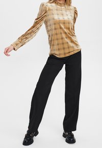 Blusa a scacchi color senape con maniche a sbuffo, colletto alto e pantaloni neri larghi. Footwear presenta mocassini neri con accenti dorati.