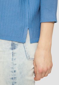Hellblau gerippter Pullover mit seitlichem Schlitz, getragen über hellen Jeans. Die Textur ist weich und gestrickt, mit 3/4-Ärmeln.