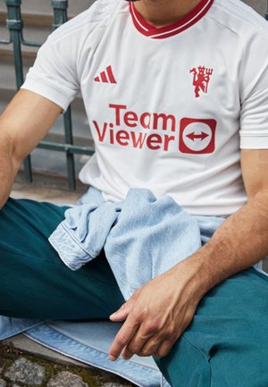 Osoba nosící bílé sportovní tričko s červeným logem TeamViewer a tyrkysové kalhoty, sedící s překříženýma nohama s světle modrou bundou na klíně.