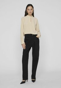 Beige långärmad blus med en nyckelhålsringning och volangärmar, kombinerad med höga svarta jeans och spetsiga svarta klackar.