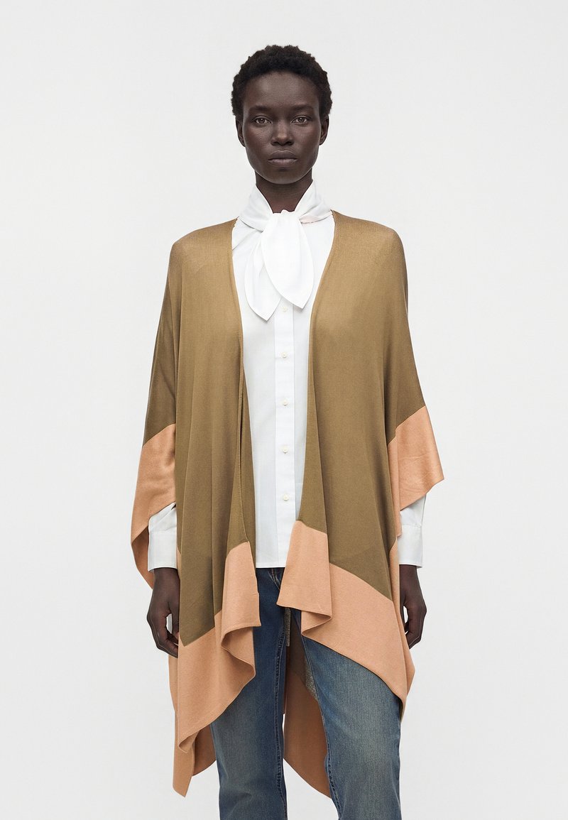 Femme noire portant un chemisier blanc avec un nœud papillon et un châle marron et pêche en blocs de couleur par-dessus un jean bleu, debout sur un fond blanc.