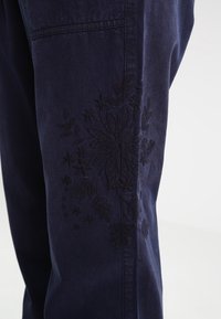 Pantalons bleu foncé ornés d'une broderie florale noire délicate sur la jambe gauche, fabriqués dans un textile robuste avec une texture lisse.