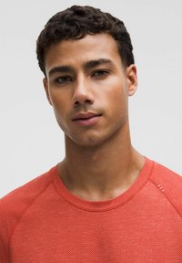 Punainen urheilupaita tekstuuripinnalla, jossa on pyöreä pääntie ja raglankädet. Olkapäässä hienovaraiset yksityiskohdat ja vartaloa myötäilevä malli.