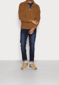 Pullover in pile marrone con zip a quarto e tasca sul petto, abbinato a jeans slim-fit blu scuro e scarpe sportive beige con accenti neri.