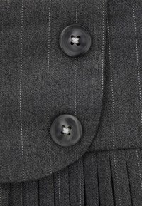 Il tessuto grigio presenta strisce verticali, due grandi bottoni neri con quattro fori e una sezione plissettata con dettagli di cucitura fine.