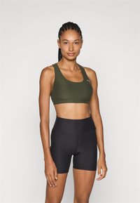 Sostén deportivo verde oliva con tirantes gruesos y un escote redondo, combinado con pantalones cortos negros de cintura alta que tienen una textura suave y costuras.