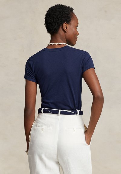 Γυναίκα με κοντά σγουρά μαλλιά που φοράει μπλε σκούρο μπλουζάκι, λευκό παντελόνι με λευκή ζώνη και κολιέ με κοχύλι, στέκεται με τα χέρια στις τσέπες.